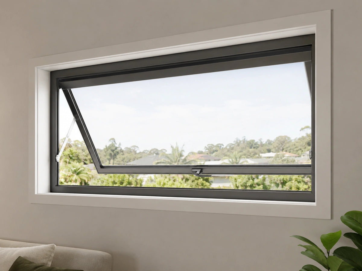 A modern black aluminum casement window with a white frame trim, shown partially open with an outdoor view of trees and sky. The window is installed on a light beige wall in a room setting with a sofa and a plant visible at the bottom corners.